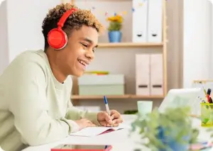 Student taking notes while attending to an online class wearing headphones